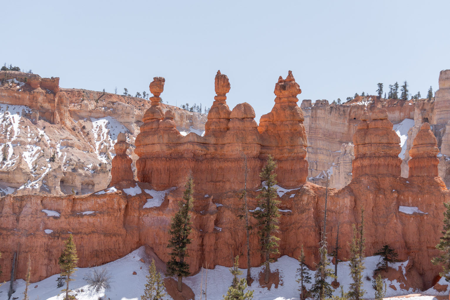 A view of some orange hoodoos that form a sort of cathedral shape against lighter hoodoos behind, light blue sky